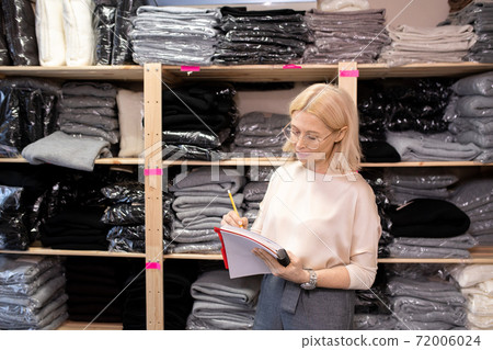 Serious mature shop assistant making notes in large copybook in storage room 72006024