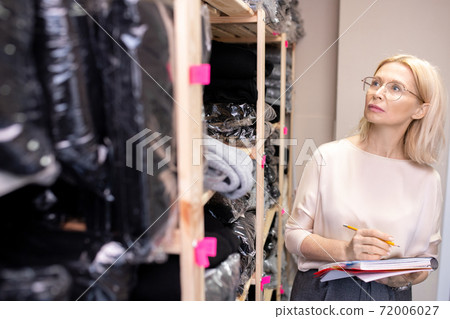 Blond mature shop assistant with pencil and copybook working in storage room 72006027
