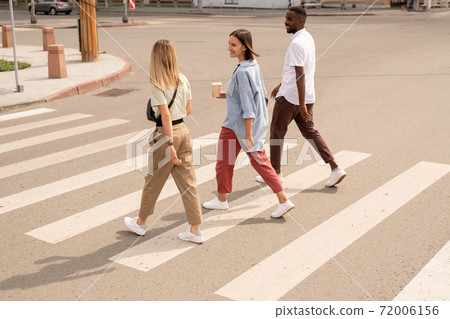 Young happy intercultural friends having drinks and talking while crossing road Young happy intercultural friends having drinks and talking while crossing road 72006156