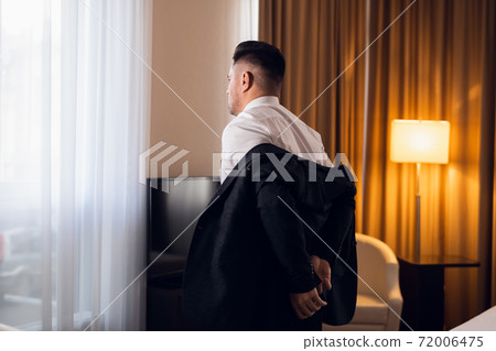 A young man taking off his jacket in a hotel room, rear view 72006475