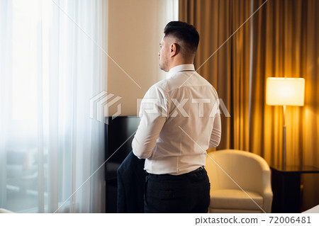 A man in a white shirt standing in front of a window in a bedroo A man in a white shirt standing in front of a window in a bedroo 72006481