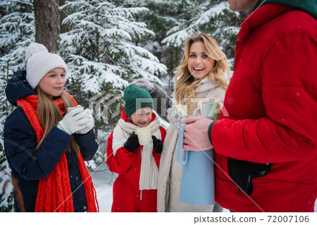 A healthy friendly family actively spends a weekend in nature in a winter spruce forest during a beautiful snowfall and enjoys hot tea from hiking mugs. A healthy friendly family actively spends a weekend in nature in a winter spruce forest during a beautiful snowfall and enjoys hot tea from hiking mugs. 72007106