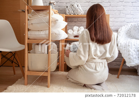 Young woman in warm robe is organizing linen closet with neatly folded towels, sheets and blankets. 72012178
