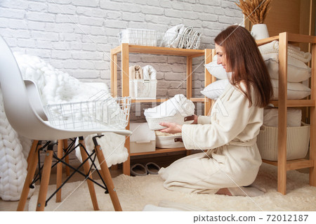 Young woman in warm robe is organizing linen closet with neatly folded towels, sheets and blankets. 72012187