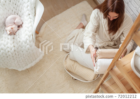 Woman in robe is sitting near wooden shelving, organizing closet by sorting and folding bath towels. Light pink colored Don Sphynx cat Woman in robe is sitting near wooden shelving, organizing closet by sorting and folding bath towels. Light pink colored Don Sphynx cat 72012212