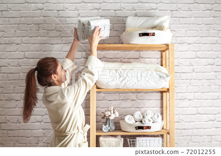 Young woman in warm robe is organizing linen closet with neatly folded towels, sheets and blankets. Young woman in warm robe is organizing linen closet with neatly folded towels, sheets and blankets. 72012255