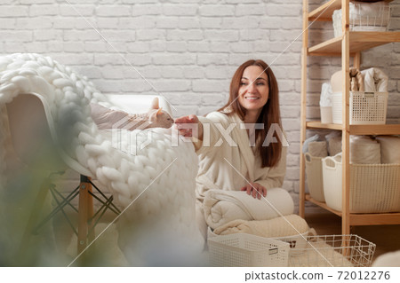 Woman in robe is sitting near wooden shelving, organizing closet by sorting and folding bath towels. Light pink colored Don Sphynx cat Woman in robe is sitting near wooden shelving, organizing closet by sorting and folding bath towels. Light pink colored Don Sphynx cat 72012276