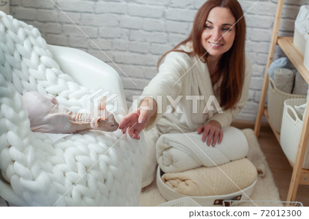 Woman in robe is sitting near wooden shelving, organizing closet by sorting and folding bath towels. Light pink colored Don Sphynx cat 72012300