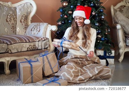 Christmas. Woman dressed white sweater Santa hat and jeans sitting on the floor near christmas tree with present box 72018677