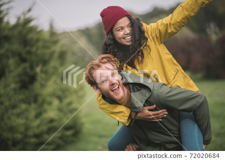 Girl in red hat sitting on her boyfriends back and smiling Girl in red hat sitting on her boyfriends back and smiling 72020684