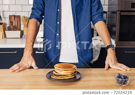 Man standing near the wooden table with a stack of freshly fried pancakes 72024219