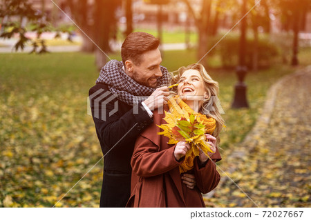 Happy young people in love, man hugging woman from behind stroking her cheek, happy couple walking in a autumn park wearing stylish coats and picking up fallen leaves. Family and people concept 72027667