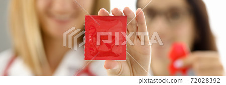 Doctors in white uniforms with red ribbon on chest hold condom in hand closeup 72028392
