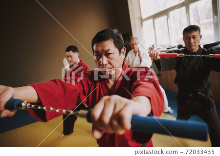 A man in a red kimono stands and holds nunchucks. 72033435