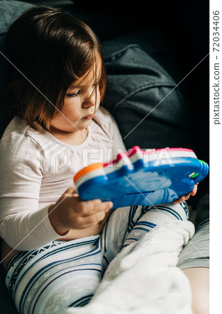 Toddler plays with colorful plastic toy at home on grey sofa. Lockdown, stay home kids.  72034406