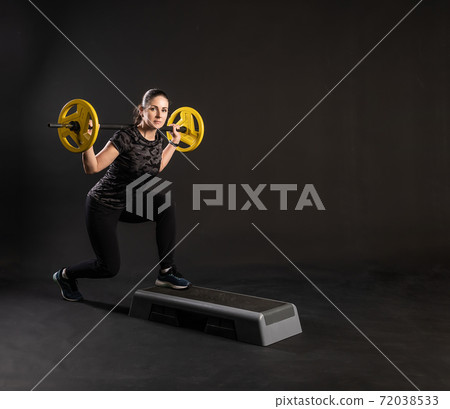 Fitness woman on a step platform, with a barbell, yellow pancakes on the bar stretches her arms. Go in for sports on a black background Cheerful 72038533