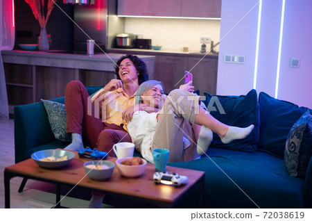 Happy boyfriend and girlfriend laughing after playing video games at home, joysticks and snacks on a table. Millennial couple enjoying spending time together. Concept of relationship. 72038619