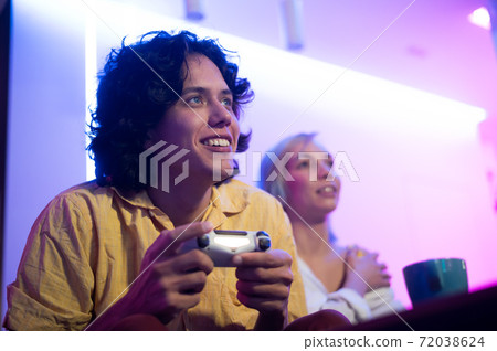 Young curly man and his girlfriend playing gaming console using joystick at home lit with neon pink and blue color. 72038624