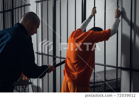 An armed guard searches a newly arrived criminal in a prison corridor against a backdrop of bars. 72039012