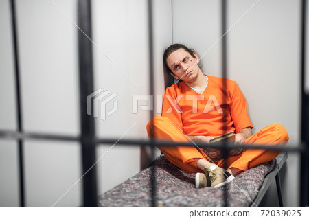 A dangerous mad criminal reads a book sitting on a bunk. In the foreground is the grid of a single cell. 72039025