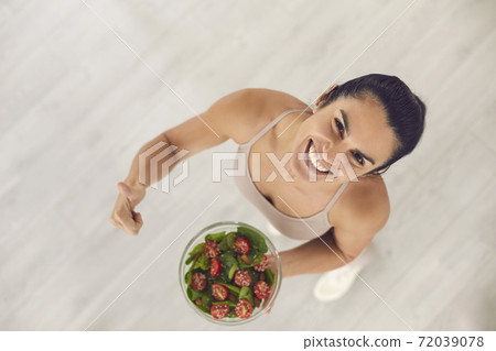 Fit woman holding bowl of vegetable salad, giving thumbs-up and smiling at camera 72039078