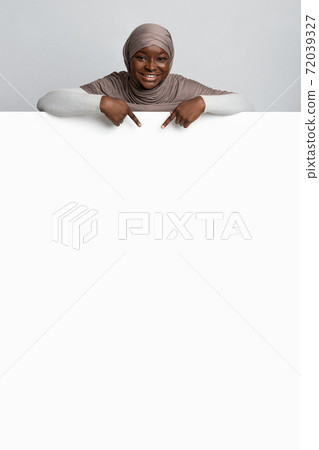 Cheerful Black Muslim Lady In Hijab Pointing At Blank White Advertisement Board Cheerful Black Muslim Lady In Hijab Pointing At Blank White Advertisement Board 72039327