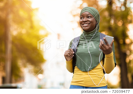 Smiling black muslim woman in headscarf walking by street and listening music on smartphone Smiling black muslim woman in headscarf walking by street and listening music on smartphone 72039477