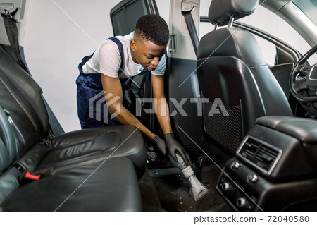 Handsome young African American man auto service worker, doing professional vacuum cleaning of salon in the car. Concept of auto service, car wash, dry and chemical wet cleaning Handsome young African American man auto service worker, doing professional vacuum cleaning of salon in the car. Concept of auto service, car wash, dry and chemical wet cleaning 72040580