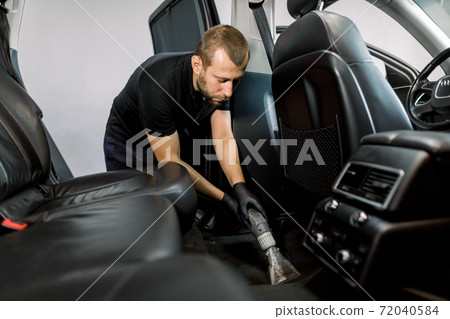 Car wash and detailing service. Male worker in black uniform and gloves, making chemical cleaning process with a vacuum cleaner and foam, releasing car floor from dust and sand 72040584