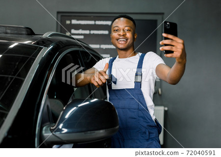 Handsome happy smiling African male worker of auto service, wearing white t-shirt and blue overalls, standing near modern black car, making selfie photo on smartphone, showing thumb up Handsome happy smiling African male worker of auto service, wearing white t-shirt and blue overalls, standing near modern black car, making selfie photo on smartphone, showing thumb up 72040591