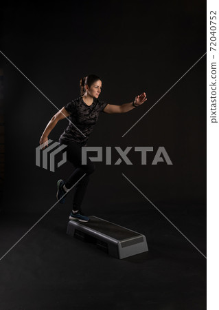Fitness girl on a step platform, pulls her leg. Go in for sports on a black background Joyful 72040752