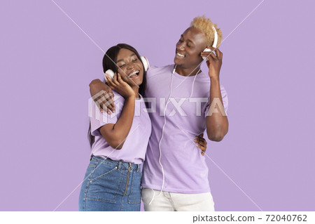 Happy black couple in headphones listening to music and hugging over lilac studio background 72040762