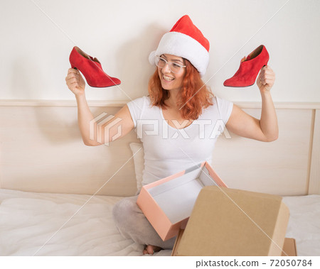 An excited woman in a Santa Claus hat takes red shoes from a cardboard box. The girl unpacks the delivered order for Christmas. Online shopping at home An excited woman in a Santa Claus hat takes red shoes from a cardboard box. The girl unpacks the delivered order for Christmas. Online shopping at home 72050784