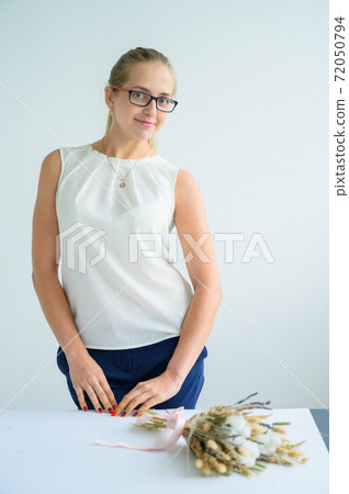 The blonde in glasses and a white blouse collects a bouquet of dried flowers and cotton in a white studio. A charming florist smiles at work. 72050794