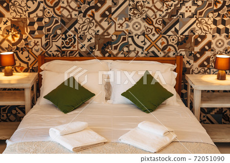 A double bed with pillows and terry towels between tables with night lamps against a wall with bright ornaments. A double bed with pillows and terry towels between tables with night lamps against a wall with bright ornaments. 72051090