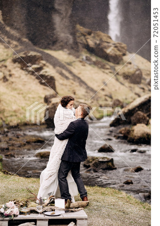 Destination Iceland wedding, near Kvernufoss waterfall. Wedding couple on the shore of a mountain river. The groom wears the bride in a woolen blanket in his hands. Destination Iceland wedding, near Kvernufoss waterfall. Wedding couple on the shore of a mountain river. The groom wears the bride in a woolen blanket in his hands. 72051453