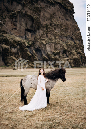 A girl in a white dress next to a white horse, with black ones. legs and mane. In a field of dry grass, against the backdrop of a cliff. Destination Iceland wedding photo session with Icelandic horses A girl in a white dress next to a white horse, with black ones. legs and mane. In a field of dry grass, against the backdrop of a cliff. Destination Iceland wedding photo session with Icelandic horses 72051490