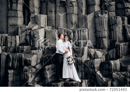 A wedding couple stands on a wall of stone pillars. The bride and groom are hugging on basalt Kekurs, on the black sandy beach of Vik. Destination Iceland wedding. A wedding couple stands on a wall of stone pillars. The bride and groom are hugging on basalt Kekurs, on the black sandy beach of Vik. Destination Iceland wedding. 72051493
