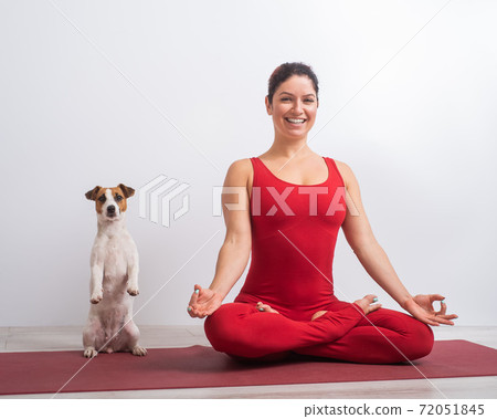 Caucasian woman in red jumpsuit sits in lotus position next to the dog. Yoga with a pet 72051845