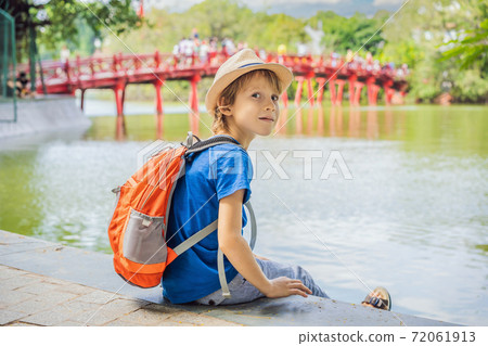 Caucasian boy tourist on background of Red Bridge in public park garden with trees and reflection in the middle of Hoan Kiem Lake in Downtown Hanoi. Traveling with children concept Caucasian boy tourist on background of Red Bridge in public park garden with trees and reflection in the middle of Hoan Kiem Lake in Downtown Hanoi. Traveling with children concept 72061913