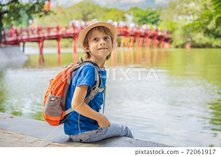Caucasian boy tourist on background of Red Bridge in public park garden with trees and reflection in the middle of Hoan Kiem Lake in Downtown Hanoi. Traveling with children concept 72061917