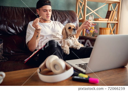 Young man sitting on sofa at home with cute dog and chatting with his friends online. Young man sitting on sofa at home with cute dog and chatting with his friends online. 72066239