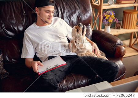 Young man sitting on sofa at home with his cute dog. Cozy office workplace, remote work, online, e-learning concept. Young man sitting on sofa at home with his cute dog. Cozy office workplace, remote work, online, e-learning concept. 72066247