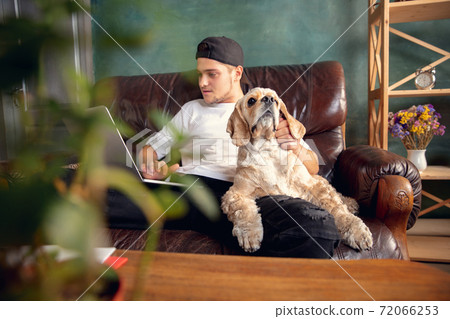 Young handsome man sitting and working at home with his cute dog. Cozy office workplace, remote work, online learning concept. Young handsome man sitting and working at home with his cute dog. Cozy office workplace, remote work, online learning concept. 72066253