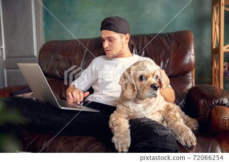 Young handsome man sitting and working at home with his cute dog. Cozy office workplace, remote work, online learning concept. Young handsome man sitting and working at home with his cute dog. Cozy office workplace, remote work, online learning concept. 72066254