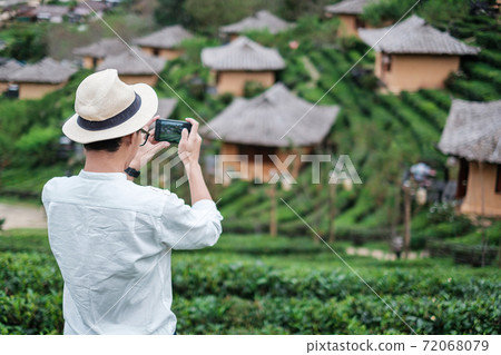 Happy tourist man in white shirt taking photo by mobile smartphone in beautiful Tea garden.Traveler visiting in Ban Rak Thai village, Mae Hong Son, Thailand. travel, vacation and holiday concept Happy tourist man in white shirt taking photo by mobile smartphone in beautiful Tea garden.Traveler visiting in Ban Rak Thai village, Mae Hong Son, Thailand. travel, vacation and holiday concept 72068079