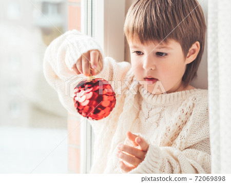 Kid with red decorative ball for Christmas tree. Boy in cable-knit oversized sweater. Cozy outfit for snuggle weather. Winter holiday spirit. New year. Kid with red decorative ball for Christmas tree. Boy in cable-knit oversized sweater. Cozy outfit for snuggle weather. Winter holiday spirit. New year. 72069898