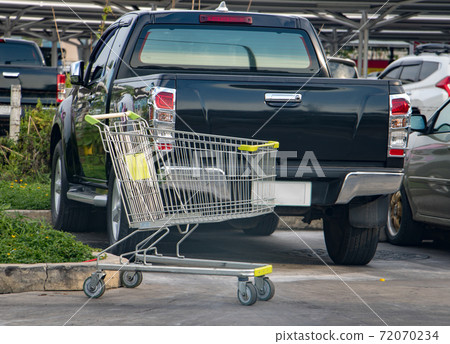 An empty shopping cart stands in a shopping mall parking lot with cars at background. 72070234