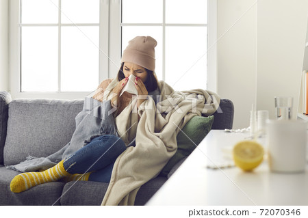 Young woman in warm clothing, socks and hat sitting on sofa, blowing nose and feeling ill Young woman in warm clothing, socks and hat sitting on sofa, blowing nose and feeling ill 72070346
