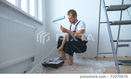 handsome man in work overalls paints the wall with a roller white paint handsome man in work overalls paints the wall with a roller white paint 72070670